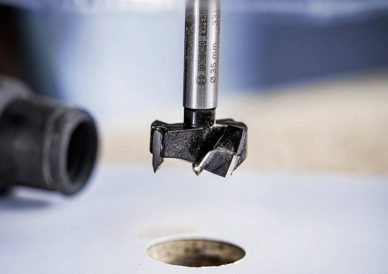 Vrták se širokými břity visí nad kulatým otvorem v prkně. Vrták je vhodný pro přesné vrtání do dřeva.