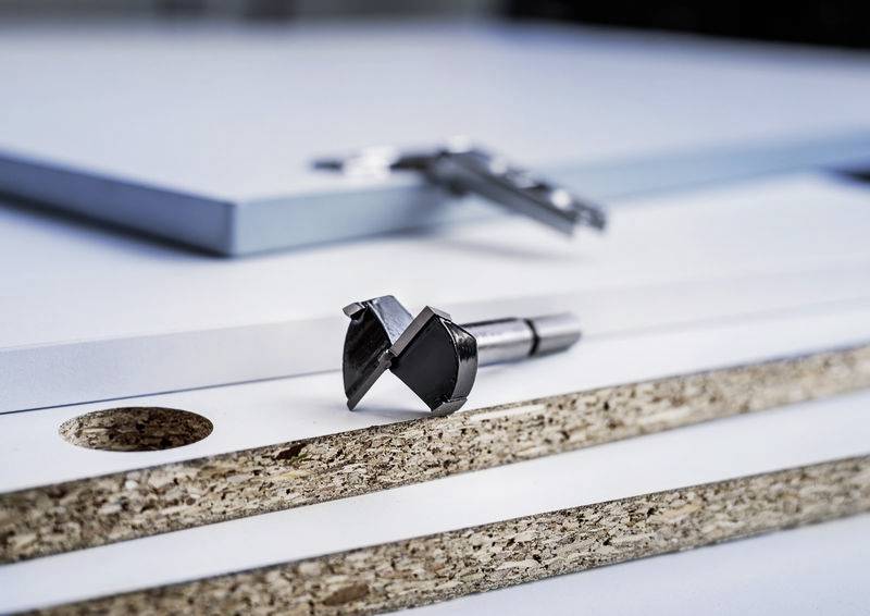 Detailní záběr sukovníku na dřevěném stole. V pozadí jsou dřevěné desky s vyvrtanými otvory. Nástroj a materiál pro práci se dřevem.