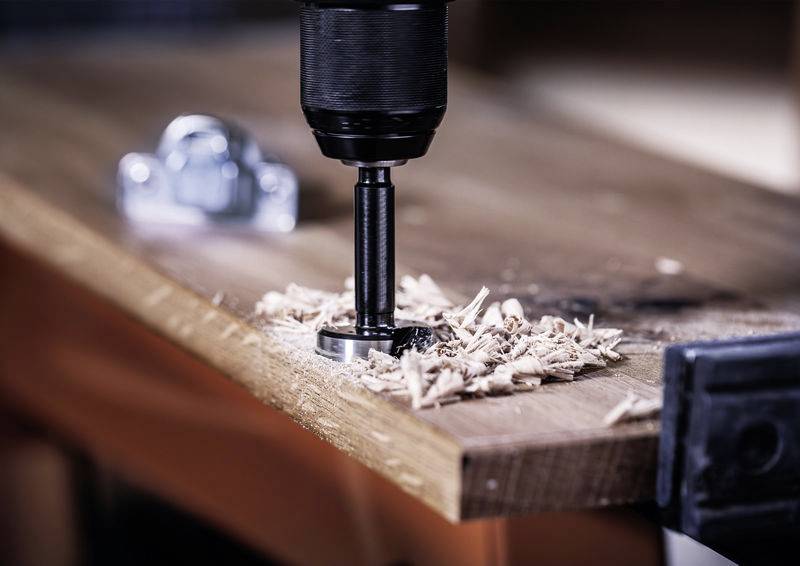 Vrtačka obrábí dřevo; vrtací hlava vytváří piliny na okraji stolu. V pozadí rozmazané nářadí.