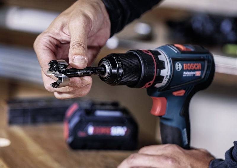 Osoba vkládá vrtací nástroj do vrtačky značky Bosch. V pozadí je vidět baterie.