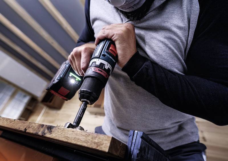 Řemeslník vrtá díru do dřevěné desky pomocí elektrické vrtačky. Je patrná jeho soustředěnost a zručnost.