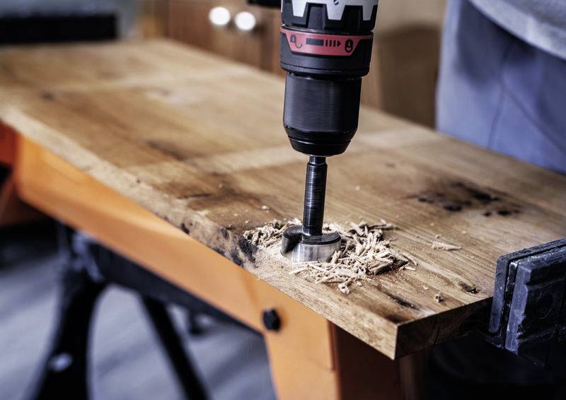 Vrtačka vrtá kulatý otvor do dřevěné desky na pracovním stole, třísky odpadávají.