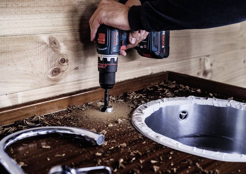 Osoba vrtá akumulátorovým vrtákem díru do dřevěné pracovní desky vedle umyvadla. Kolem jsou roztroušené dřevěné třísky.