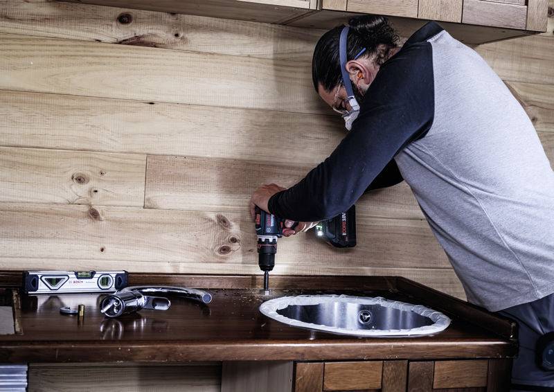 Osoba instaluje pracovní desku vedle umyvadla a používá vrtačku. Pracovní nástroje leží vedle ní.