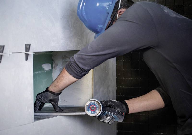 Osoba nosí modrý bezpečnostní helmu a černé rukavice. Používá elektrické nářadí k rozšíření díry ve zdi.