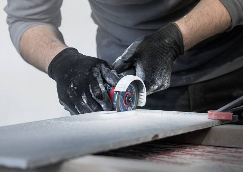 Osoba pracuje v rukavicích u brusky při opracování tvrdé desky. Během broušení vzniká prach.