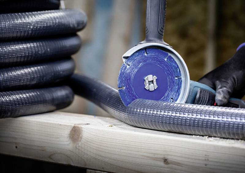 Člověk řeže kovovou trubku úhlovou bruskou, která leží na dřevěném stole. Pozadí je rozmazané.