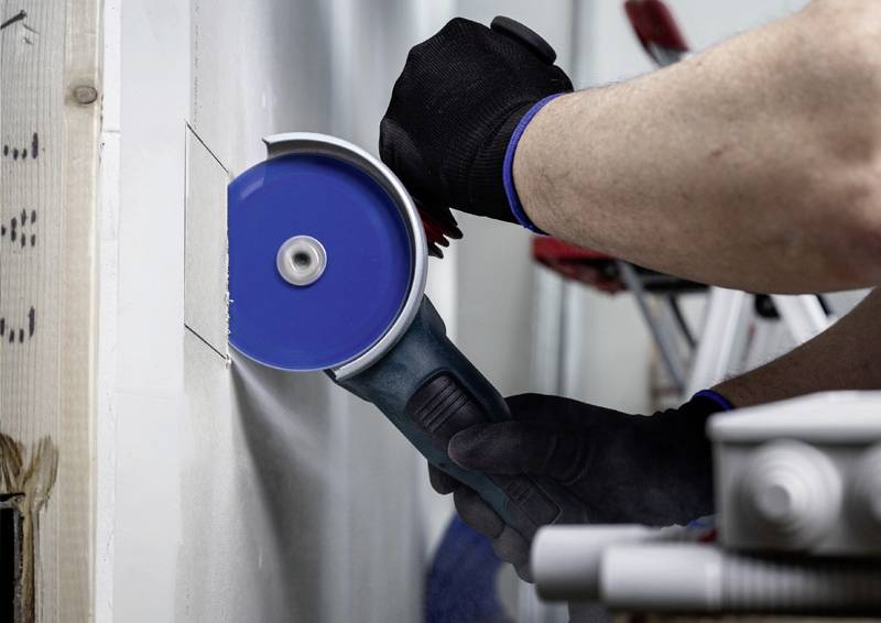 Osoba řeže modrou kotoučovou pilou díru do bílé zdi. Má na sobě černé rukavice.