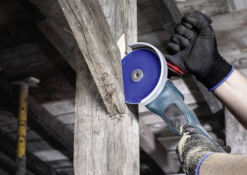 Řemeslník používá úhlovou brusku k opracování dřevěného trámu. Má nasazené ochranné rukavice a pracuje v dřevěné konstrukci.