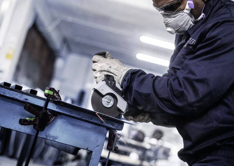 Dělník v ochranných brýlích a respirátoru brousí kov v dílně. V pozadí silné světlo.