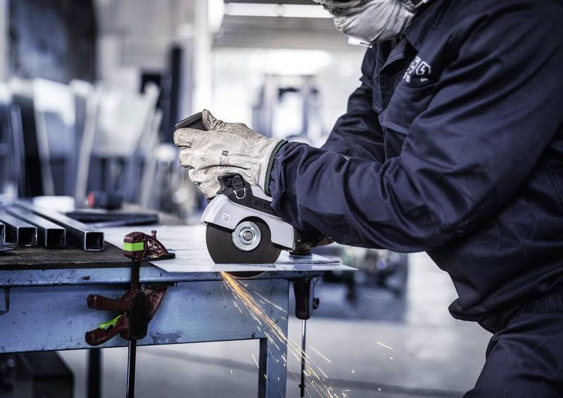 Osoba v pracovním oděvu řeže úhlovou bruskou kov na stole. Při práci odlétávají jiskry.