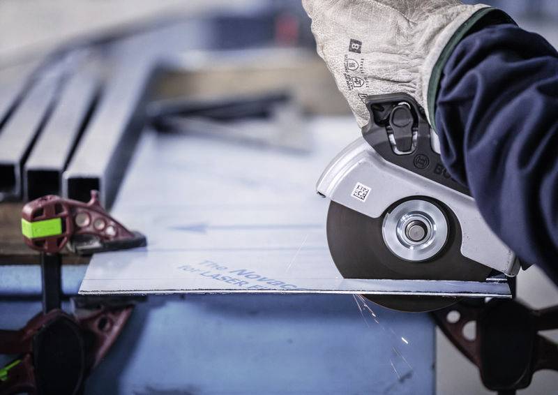 Člověk řeže úhlovou bruskou kovový díl. Je oblečen v ochranném oděvu. Od kovu odlétávají jiskry.