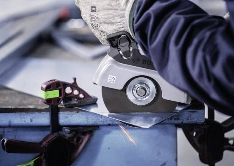 Elektrická bruska obrábí kovovou desku na stole, přičemž z povrchu odlétávají jiskry. Svorka upevňuje materiál.