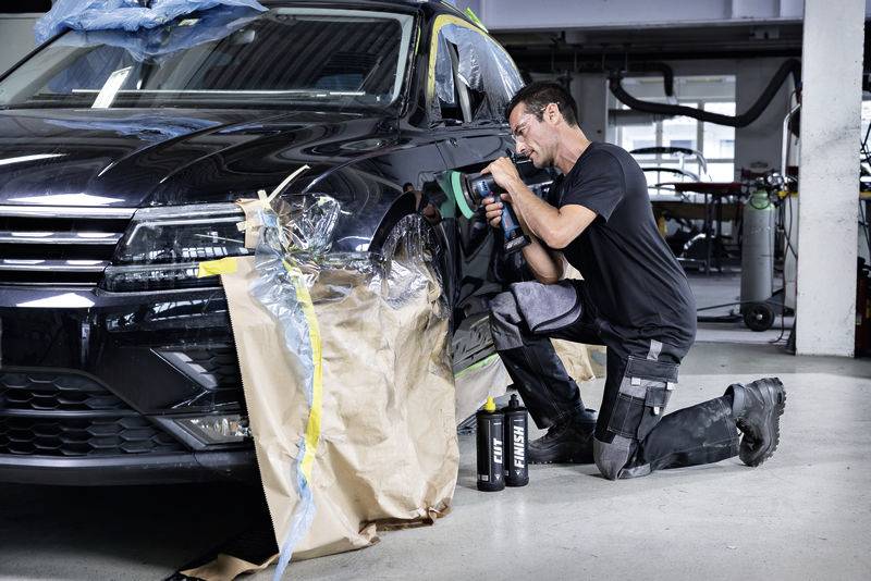 Muž klečí a leští přední levou část černého automobilu pomocí zařízení; situace opravy nebo údržby v autoservisu.