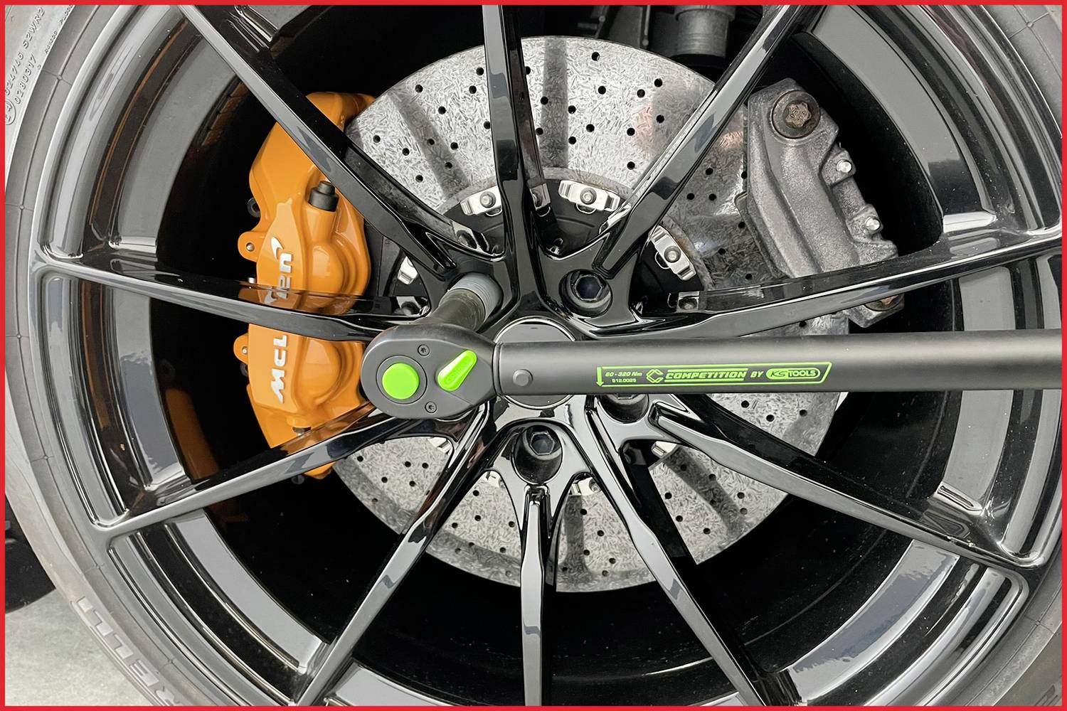 Detailní záběr pneumatiky automobilu s černým diskem a oranžovým brzdovým třmenem. Na kole je připevněn momentový klíč.