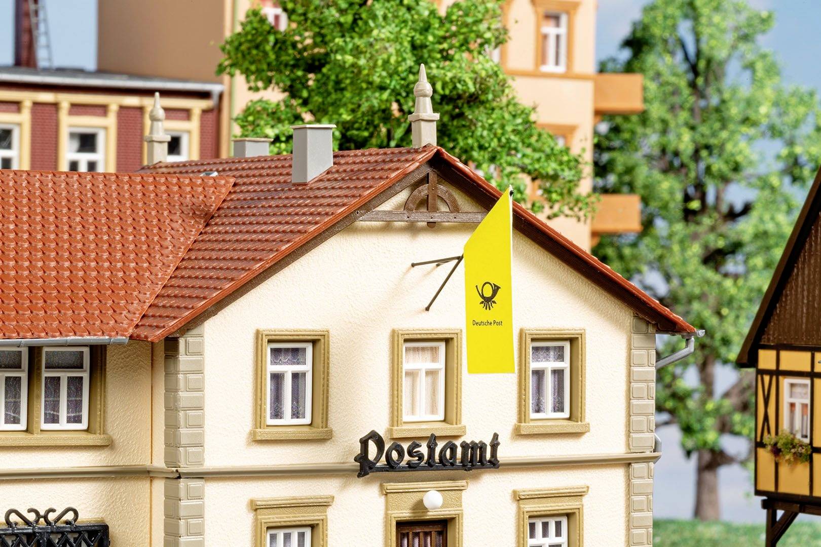 Budova s nápisem 'Pošta', žlutá vlajka s logem poštovního rohu a stromy v pozadí.