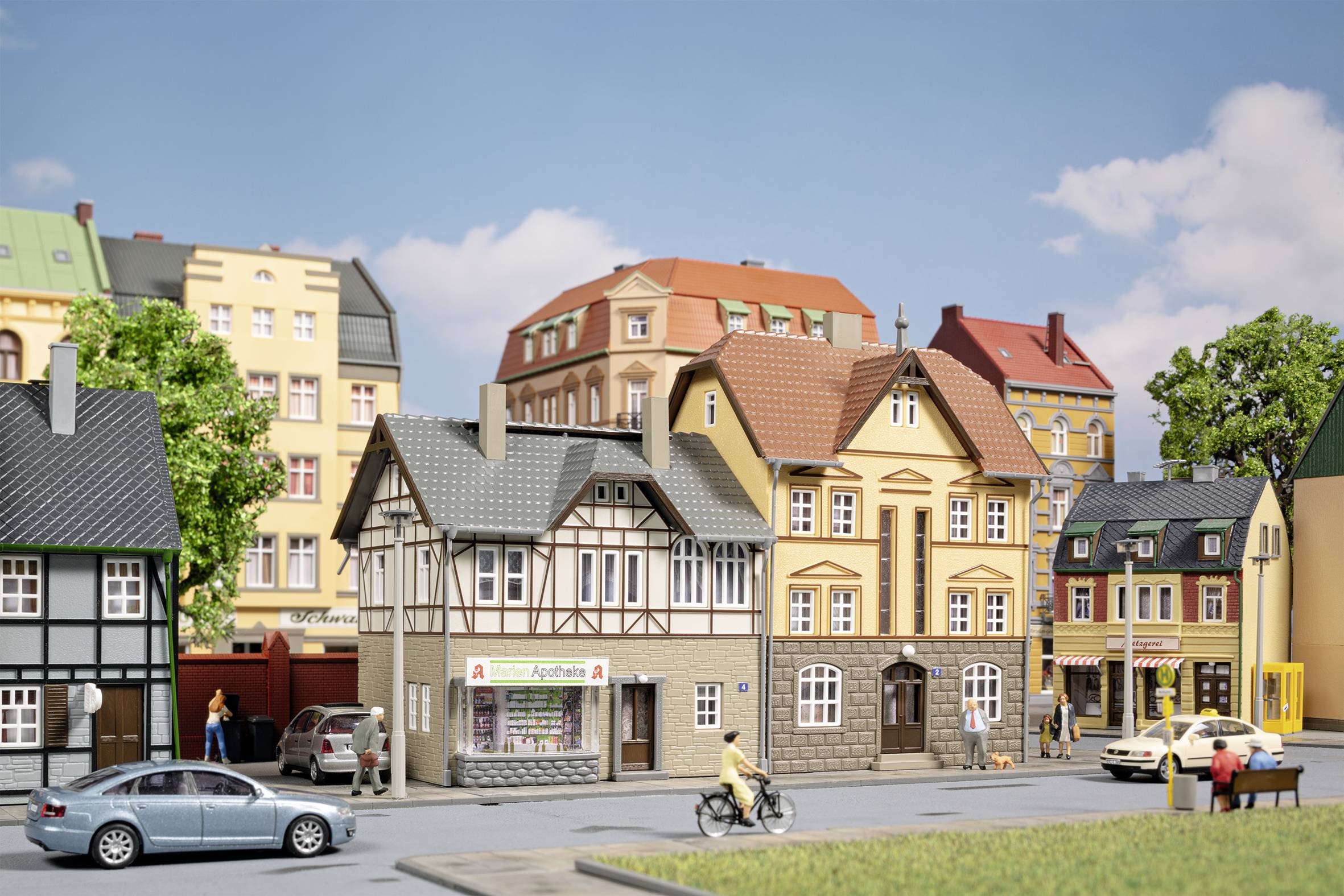 Pohled na město s modely domů v tradičním stylu, osobou na kole a auty na silnici před modrým nebem.
