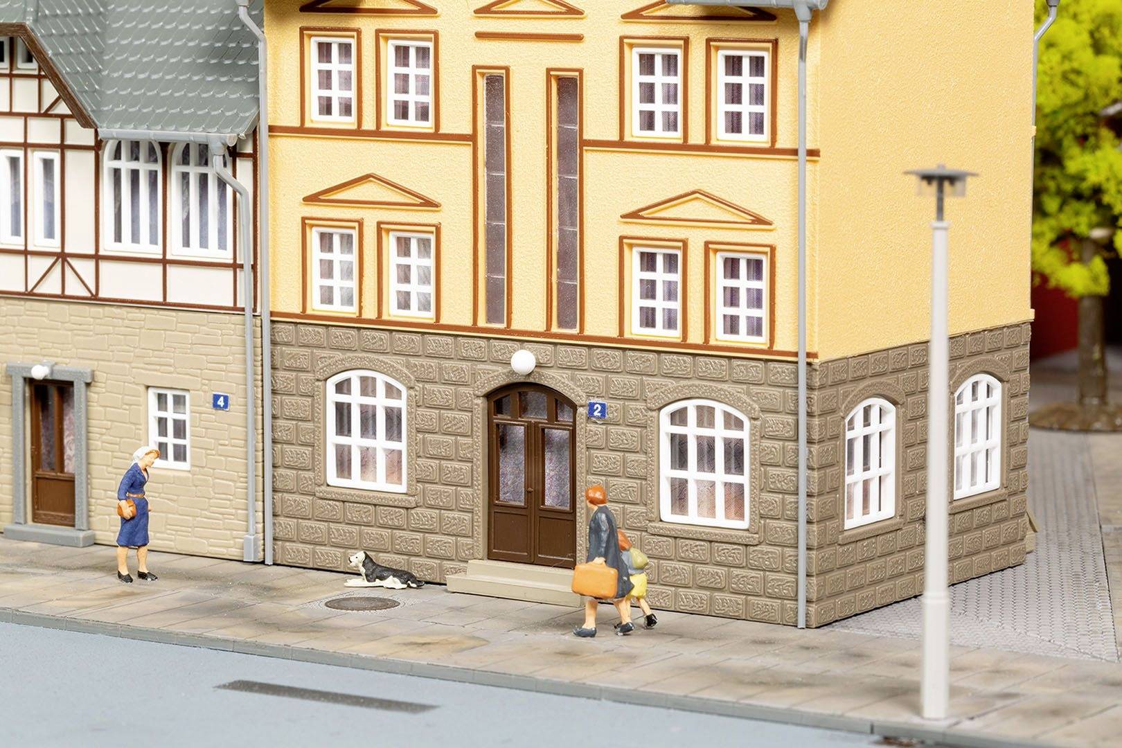 Pouliční scéna s modelářským domem. Jedna osoba s taškou prochází kolem, další stojí a kočka leží na chodníku.