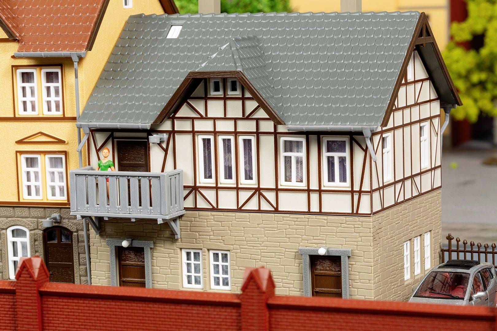 Miniaturní model hrázděného domu se dvěma patry, balkónem a šedou střechou. V popředí červený plot.