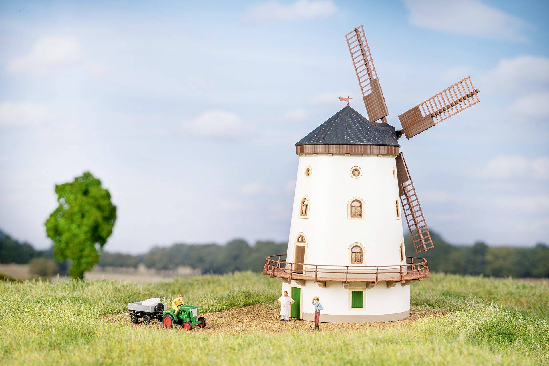 Model větrného mlýna s otáčivými křídly stojí na poli. V popředí je malý traktor s přívěsem.