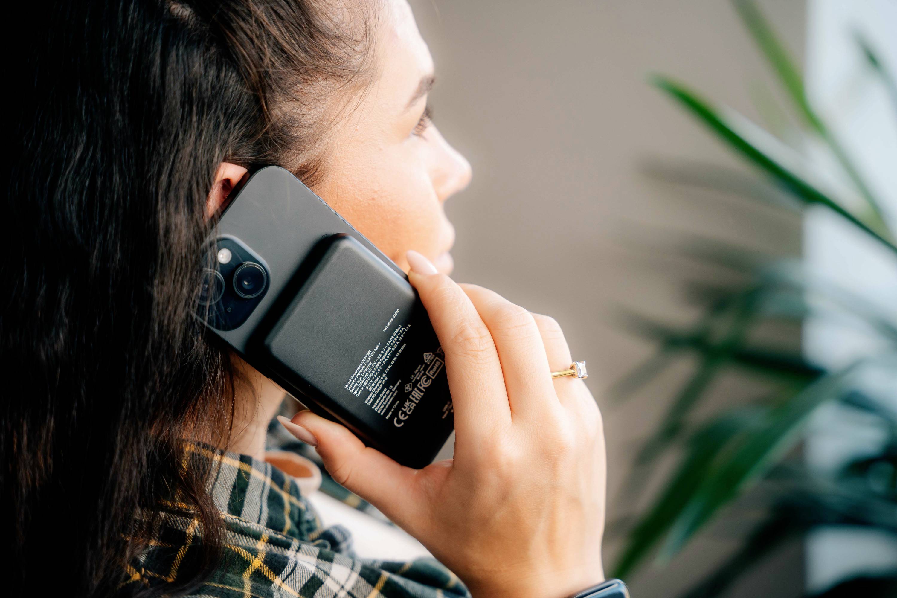 Osoba telefonuje pomocí mobilního telefonu přiloženého k uchu, pozadí je rozmazané a je vidět rostlina.