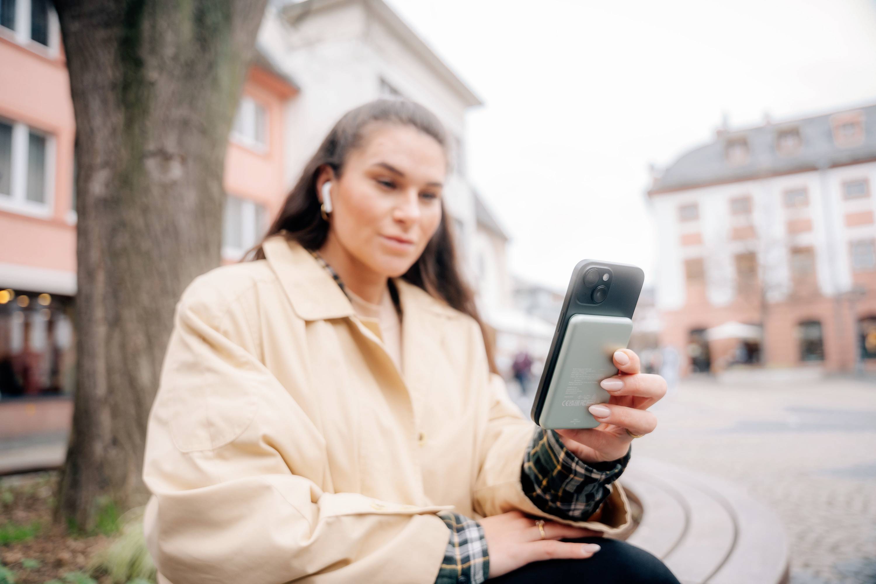 Žena sedí na lavičce ve městě a dívá se do svého smartphonu, má světlou bundu a sluchátka, obklopena budovami v pozadí.