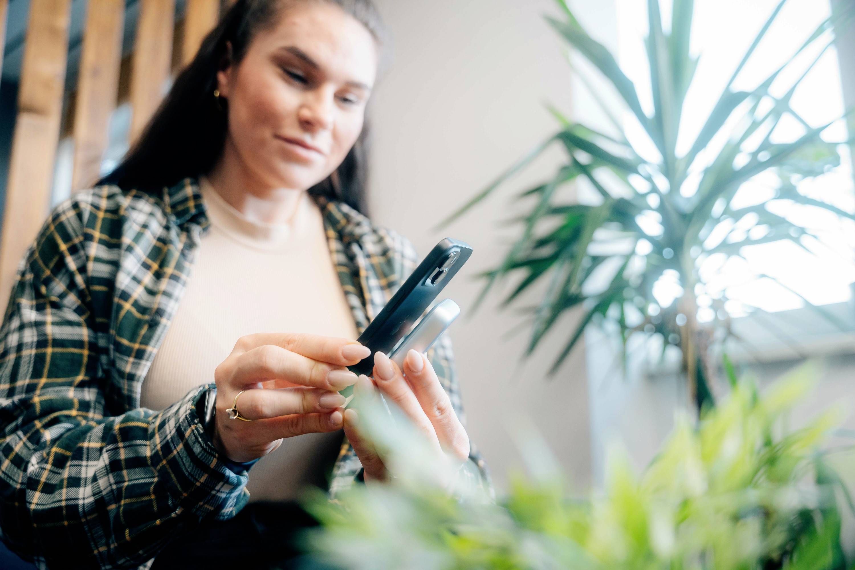 Osoba se dívá na smartphone v interiéru s rostlinami v pozadí. Má na sobě kostkovanou košili.