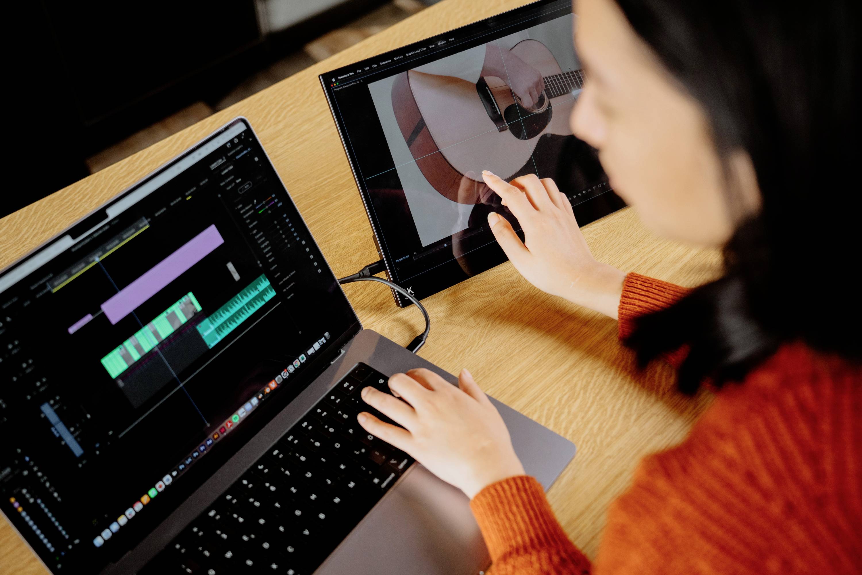 Jedna osoba pracuje na laptopU a tabletu. Na laptopU je vidět videoeditor s audio stopami, zatímco tablet zobrazuje obrázek kytary.
