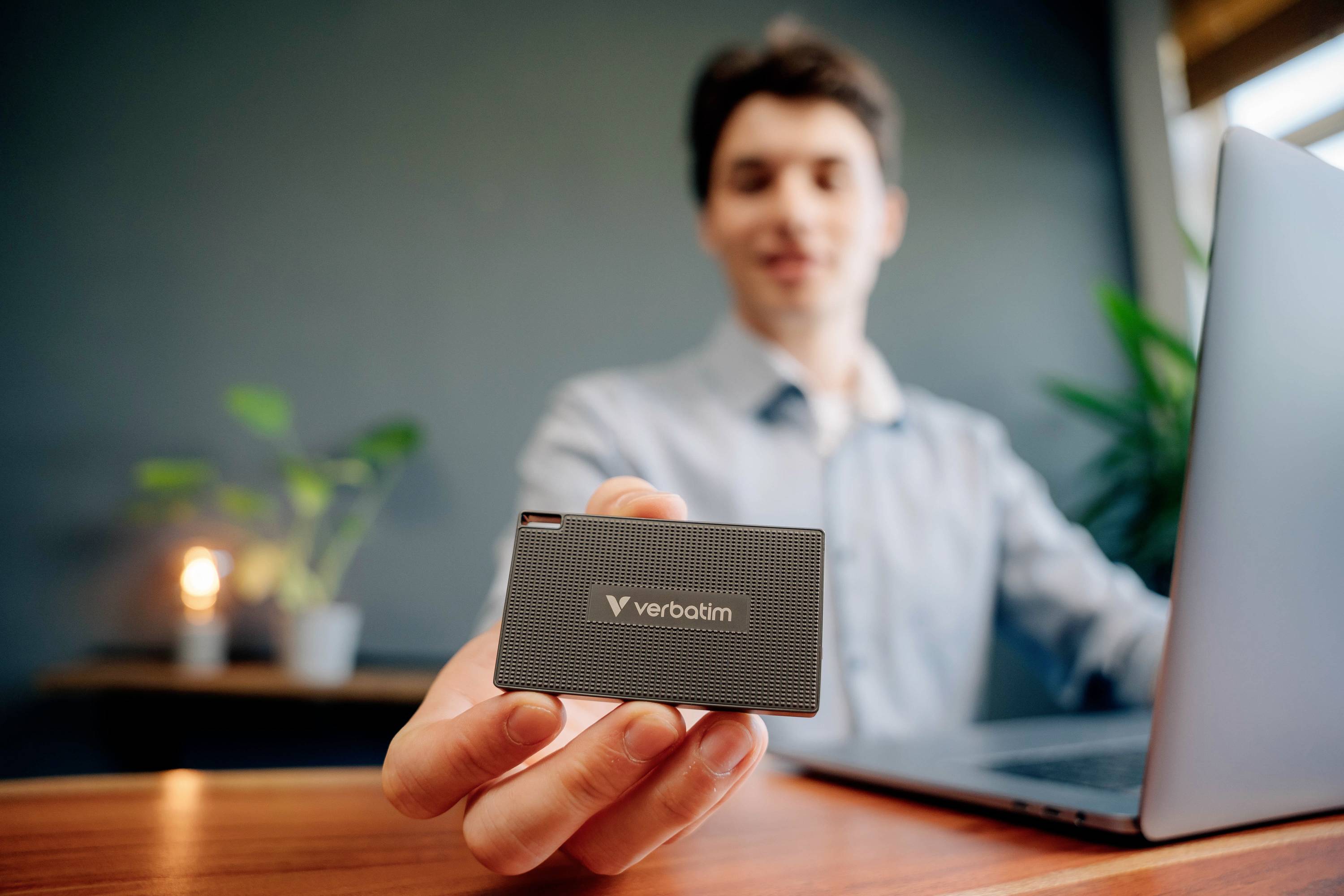 Muž sedí u laptopu a drží externí disk značky Verbatim před kamerou. V pozadí jsou rostliny.