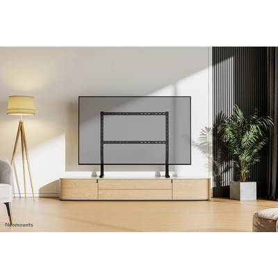 Stojan na televizi před televizí v moderním, minimalisticky zařízeném obývacím pokoji s lampou a rostlinou v pozadí.