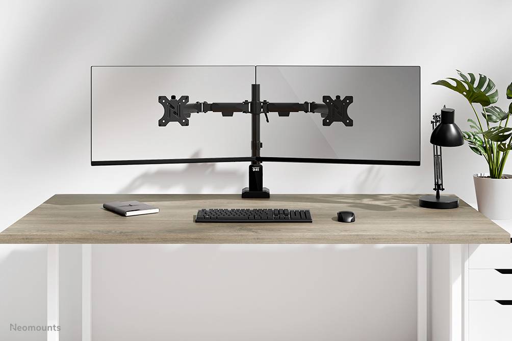 Dva monitory na psacím stole s klávesnicí a myší, vlevo notes, vpravo lampa a rostliny v pozadí.