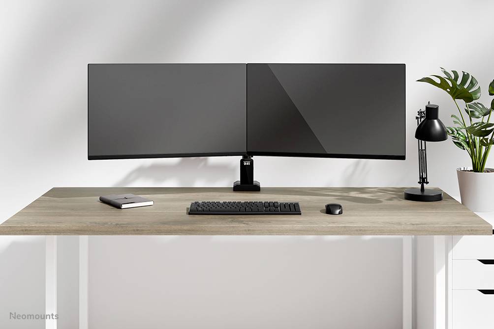 Moderní psací stůl se dvěma monitory, klávesnicí, myší a stolní lampou. Vedle je umístěna rostlina.
