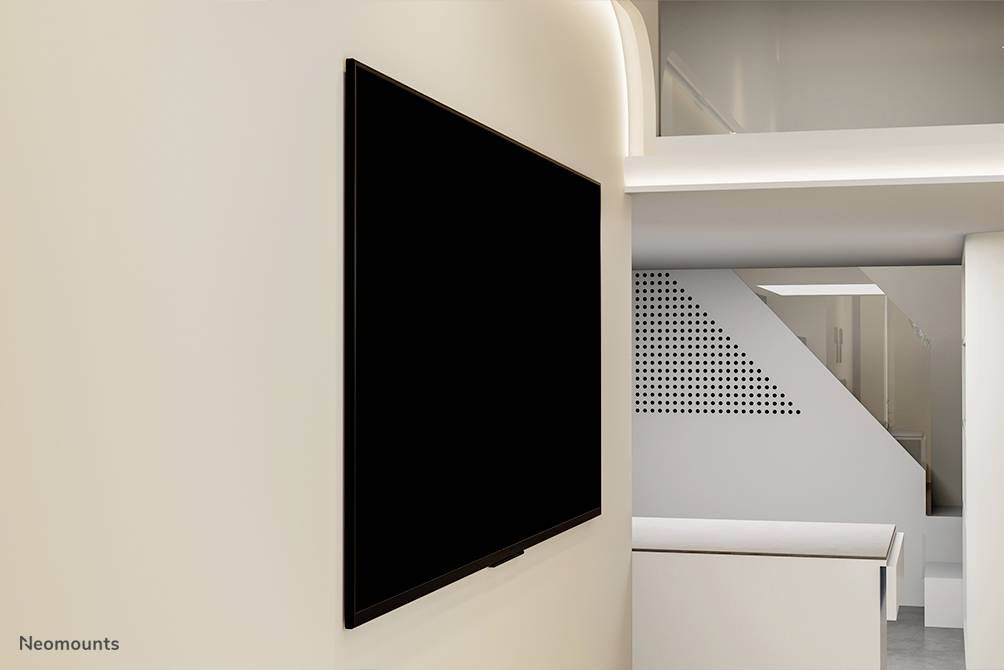 Na světlé stěně visí velký černý plochý displej. V pozadí jsou patrné moderní architektonické detaily.