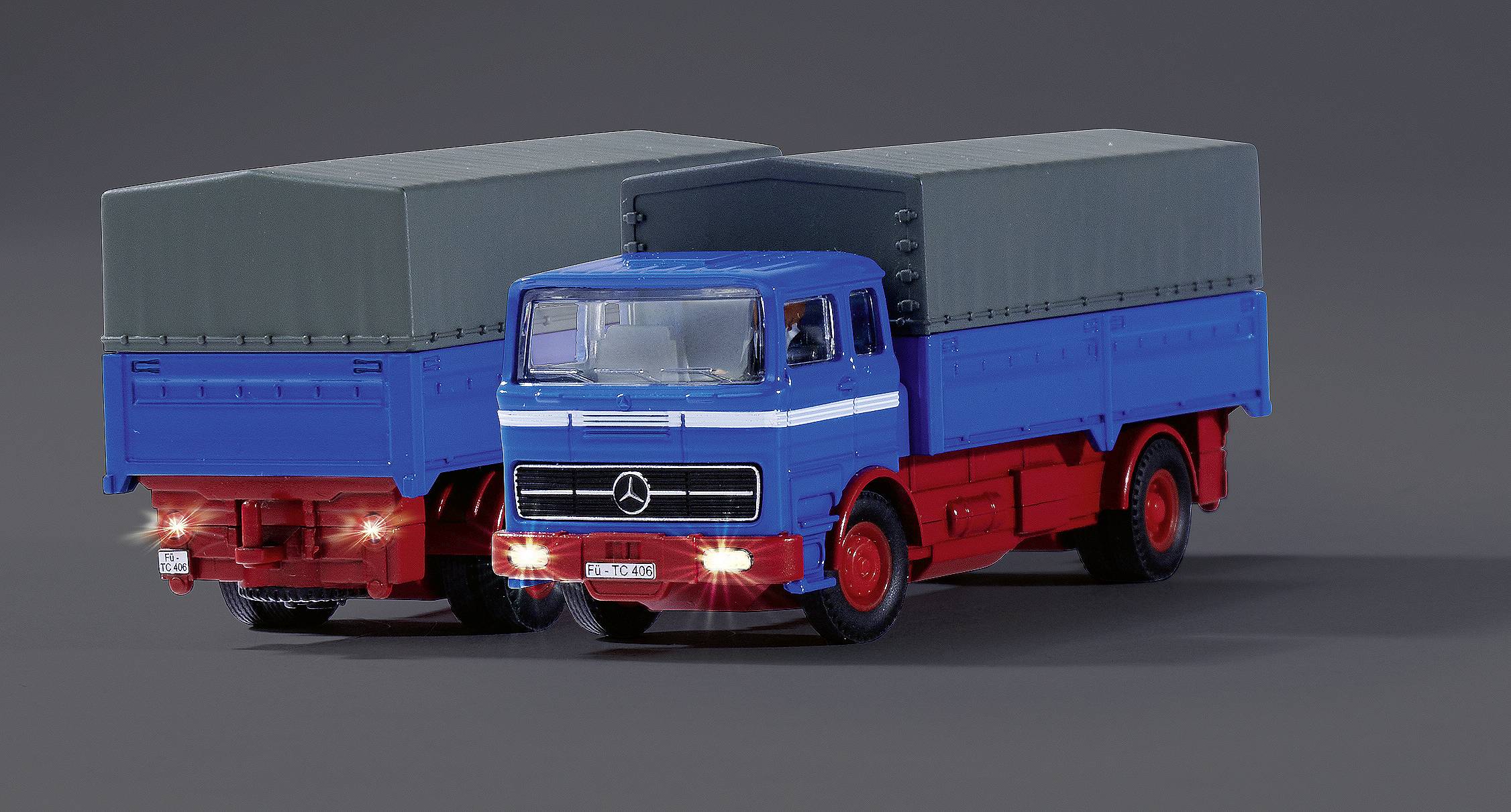 Dva modelové nákladní automobily s modrými kabinami řidiče a červenými nástavbami na šedém pozadí. Oba mají šedé plachty.