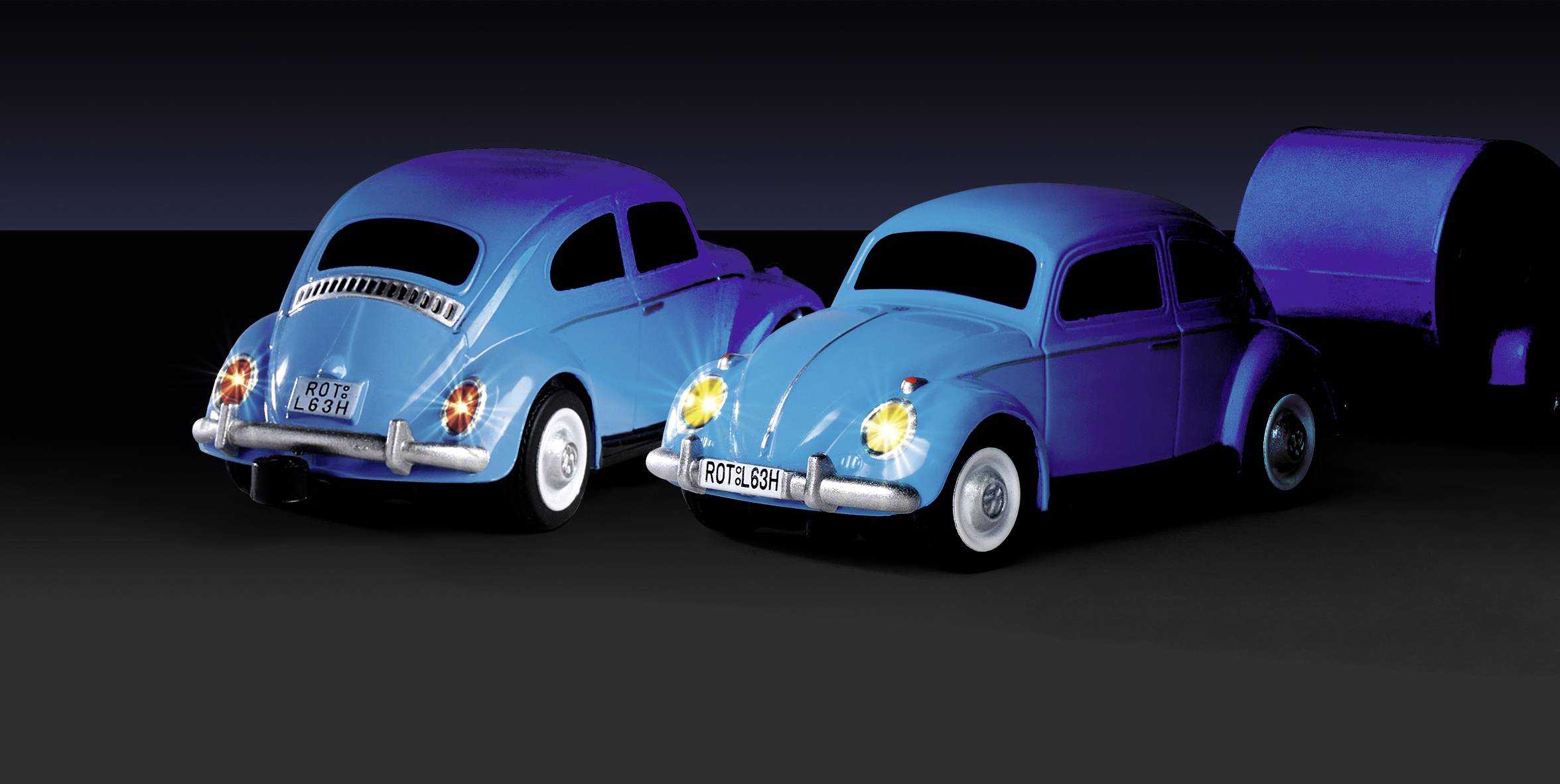 Dvě modré hračkové autíčka podobné volkswagenu broukovi stojící vedle sebe. V pozadí přívěs.