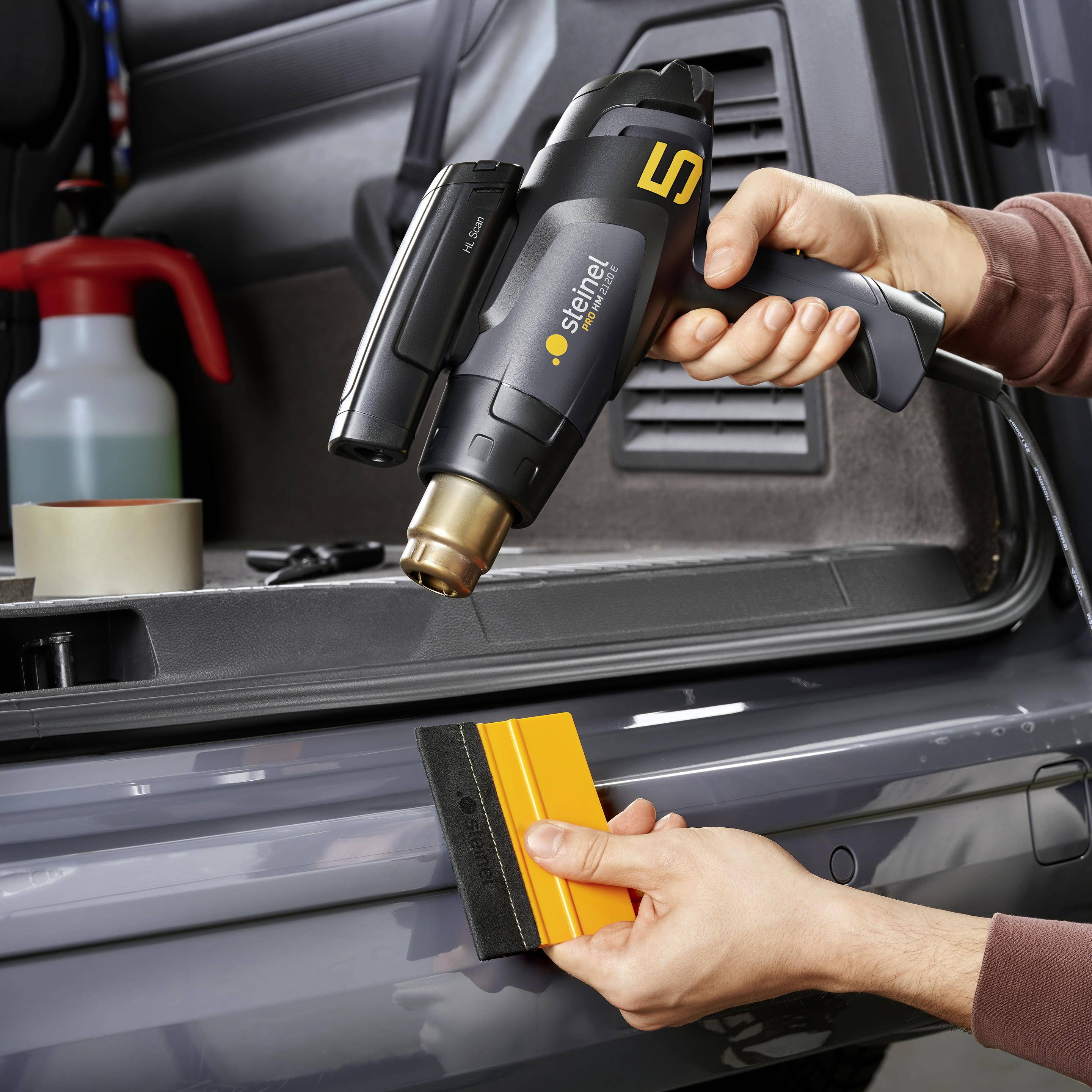 Dva páry rukou používají horkovzdušnou pistoli k nanášení fólie na lak automobilu. Nástroj a kapalina jsou v pozadí.