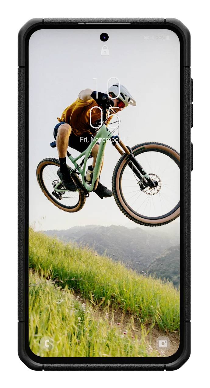 Cyklista ve vzduchu, má na sobě helmu a oranžové oblečení, jede přes travnaté kopce. Na obrazovce smartphonu je viditelný čas.