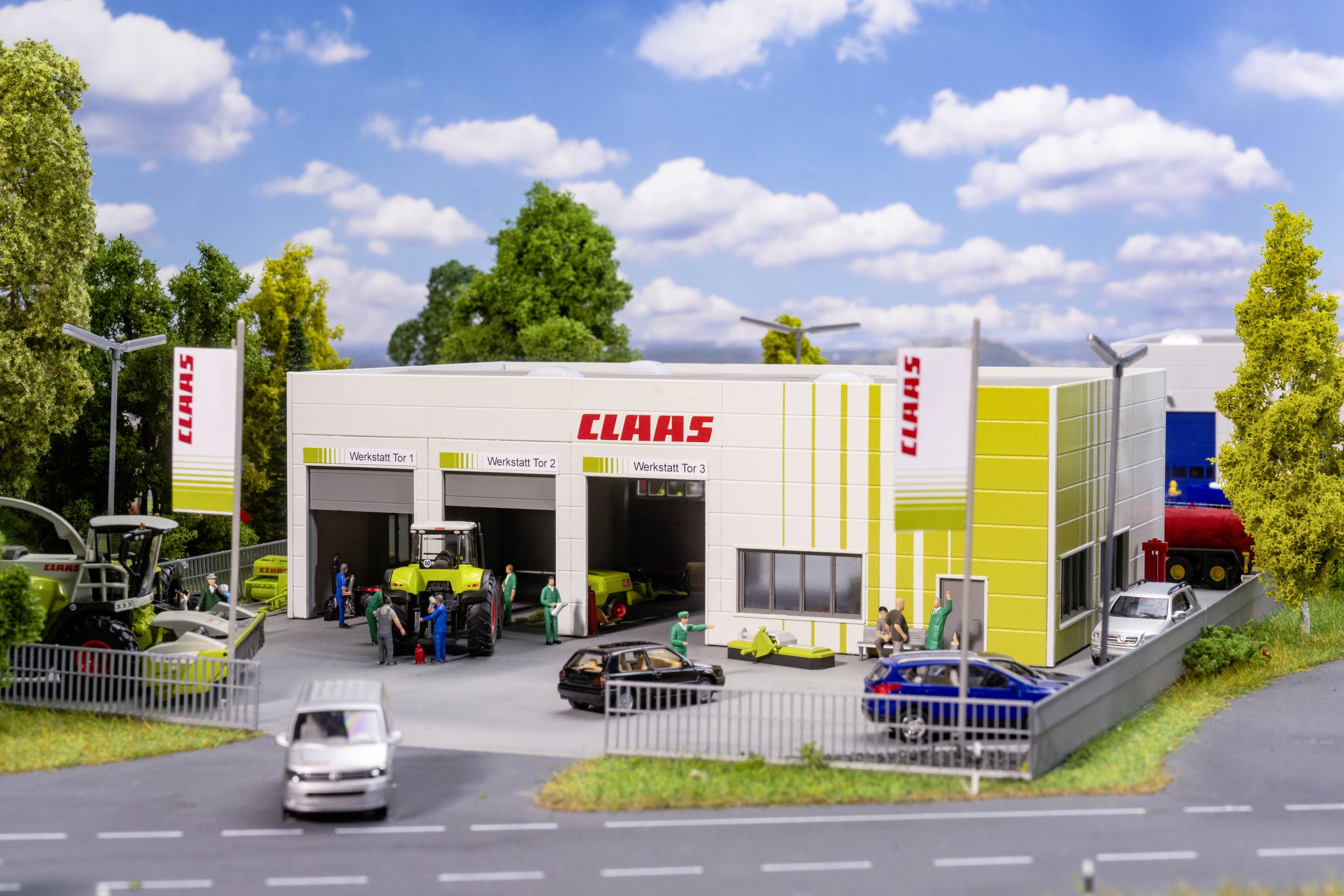Miniaturní model dílny CLAAS s traktory a auty před garáží. Několik figurek je vzájemně propojeno v interakci.