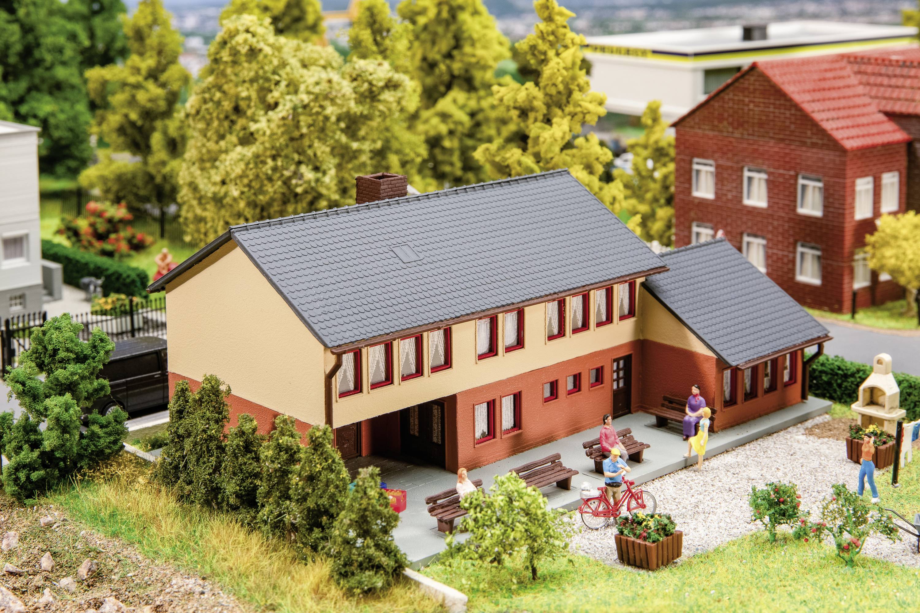 Miniaturní model ukazuje budovu s červenou taškovou střechou, obklopenou stromy a figurkami v kulise vesnice.