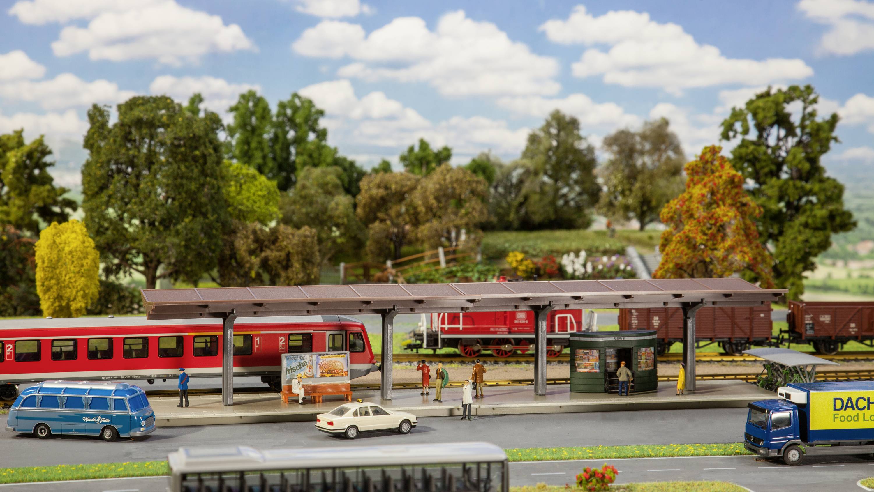 Miniaturní nádraží s červeným vlakem, obklopené stromy a silnicemi, ukazuje lidi, kteří nastupují a vystupují.