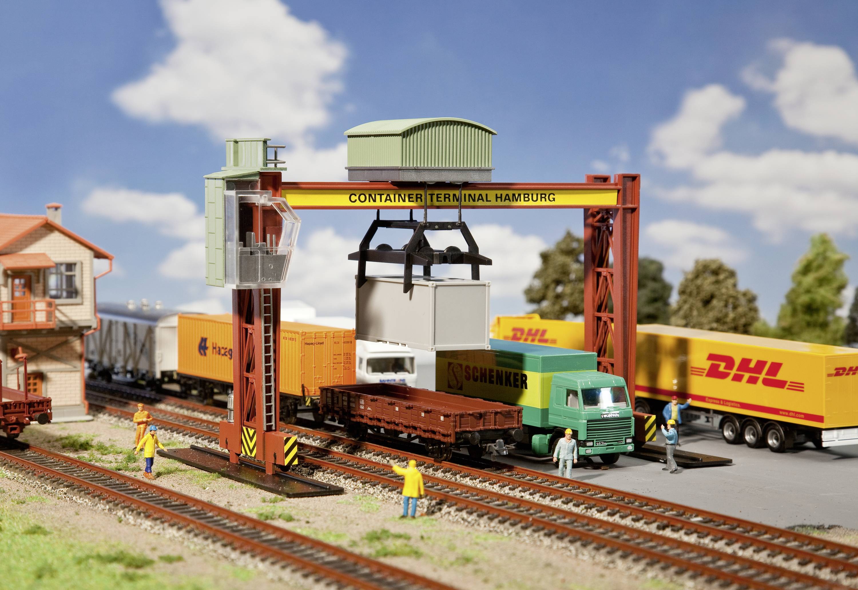 Miniaturní kontejnerový terminál zobrazuje vysokozdvižný jeřáb, který nakládá kontejner na vlak. V pozadí jsou nákladní vozy DHL.