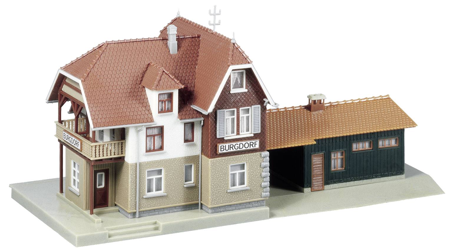 Model malé nádražní budovy s červenou cihlovou střechou, béžovými stěnami a přístavbou z tmavého dřeva.