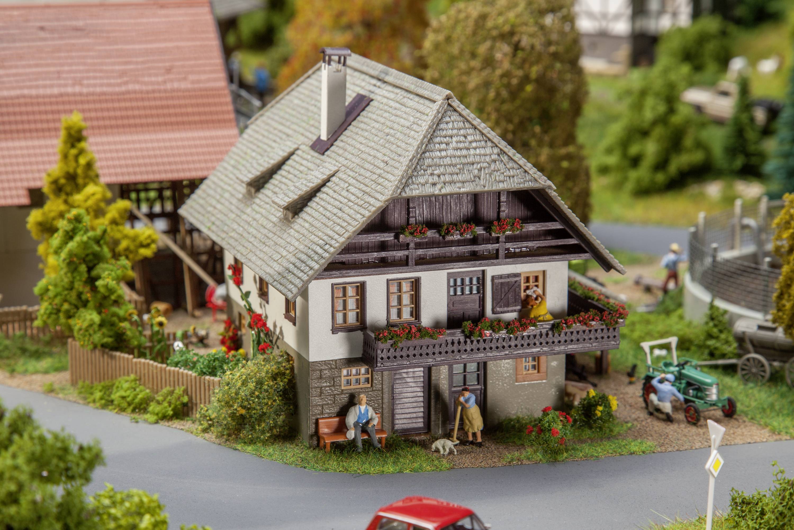 Model dvoupatrového domu s květinami na balkóně, vedle figurky lidí a vozidel v rurální scenérii.