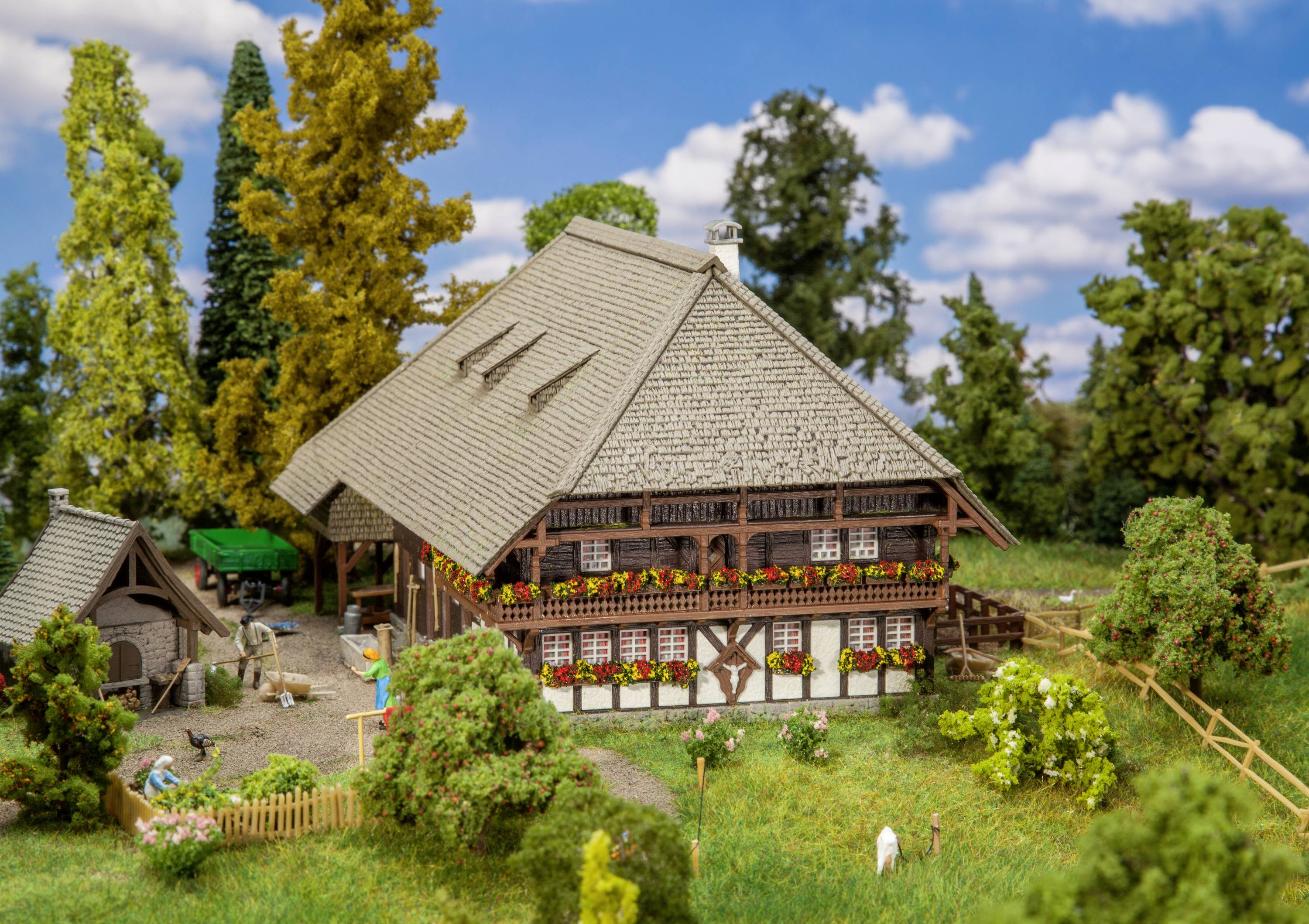 Tradiční hrázděný dům v rurálním prostředí s květinovou výzdobou na balkónech, obklopený stromy a malým dvorkem v popředí.