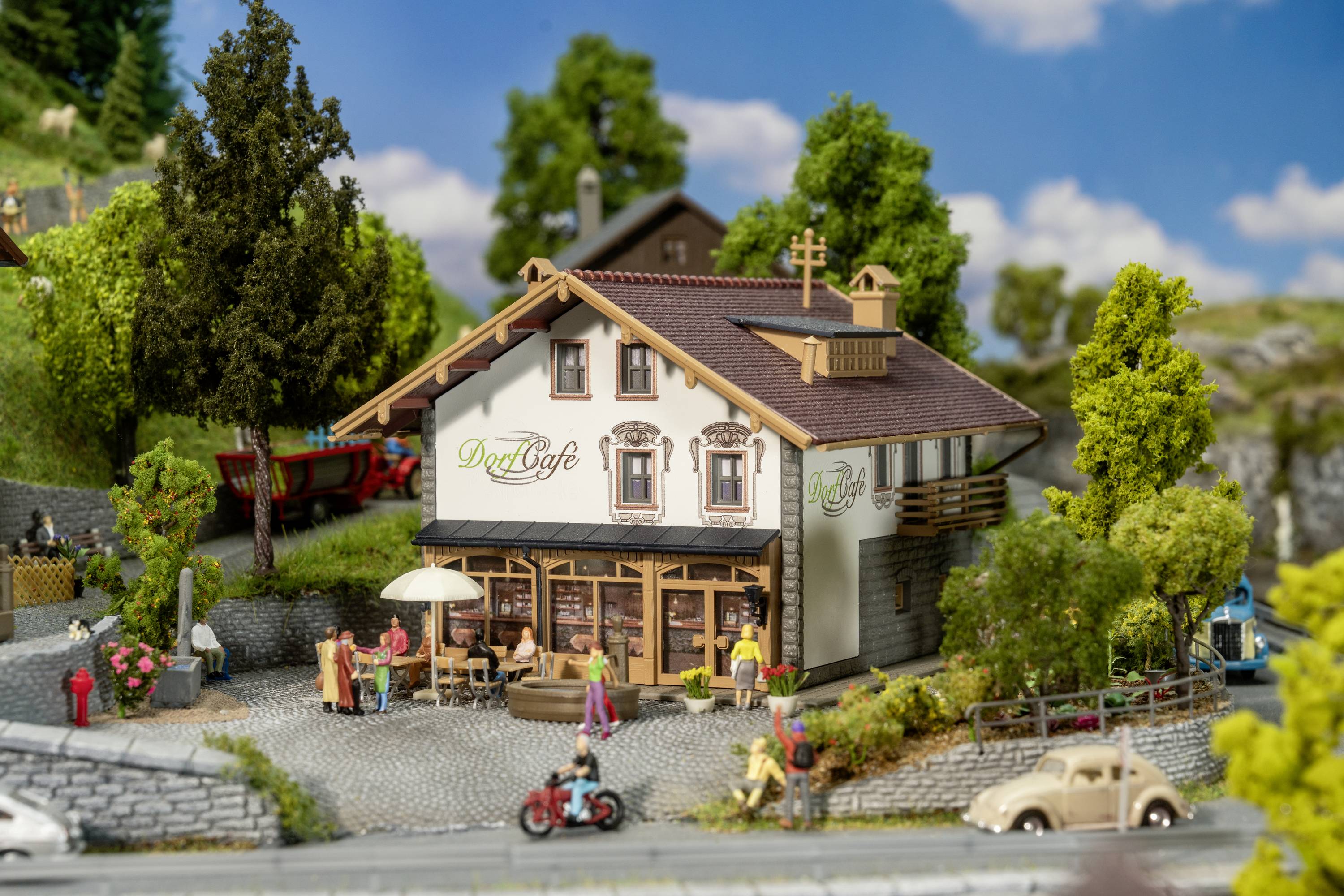 Miniaturní model kavárny nazvané 'Vesnická kavárna', obklopený stromy, květinami a miniaturními figurkami. Scéna v rurálním prostředí.
