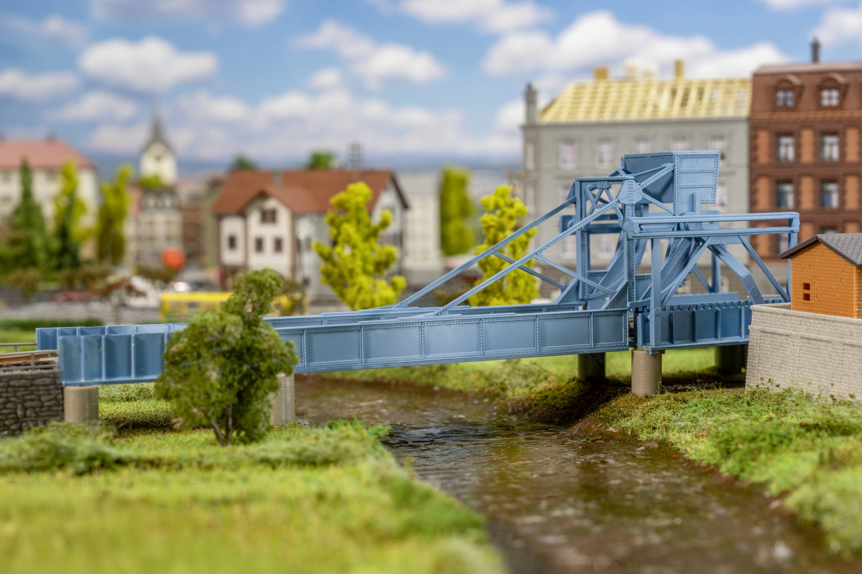 Modrý model pohyblivého mostu přes řeku, obklopený miniaturními domy a stromy, s oblačnou oblohou v pozadí.
