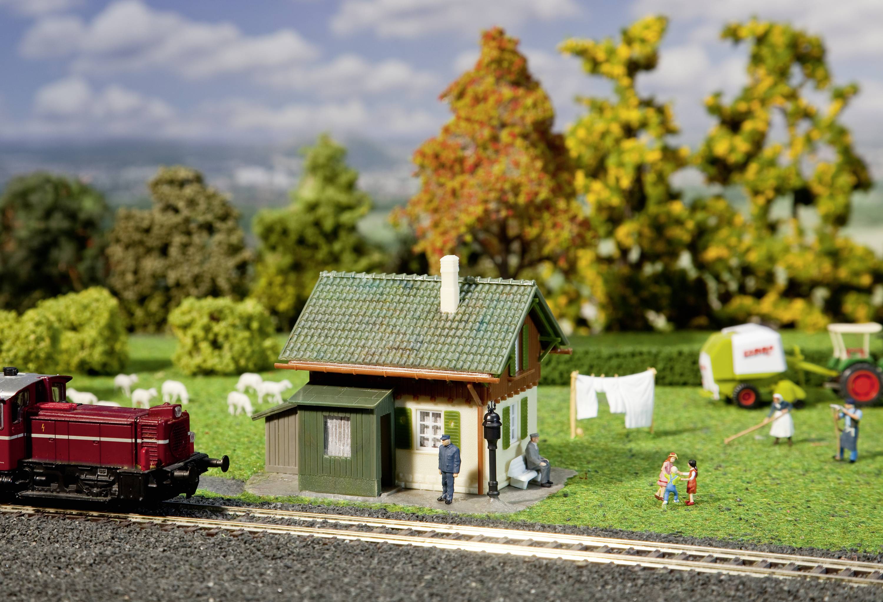 Miniaturní scéna s malým domkem a prádlem věšeným na šňůře, obklopená stromy. V popředí vede modelová železniční trať.