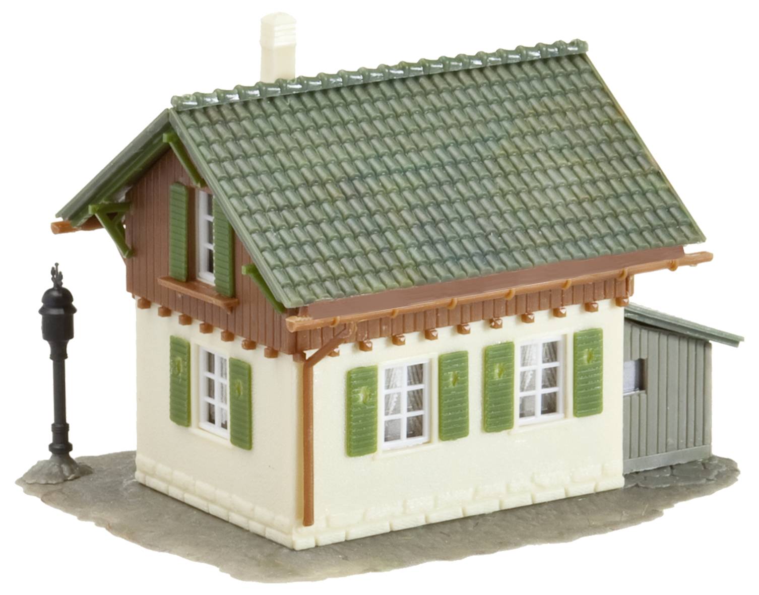 Malý modelový dům se zelenou střechou, zelenými okenicemi, hnědými dřevěnými detaily a přilehlou stodolou; lampa vlevo.