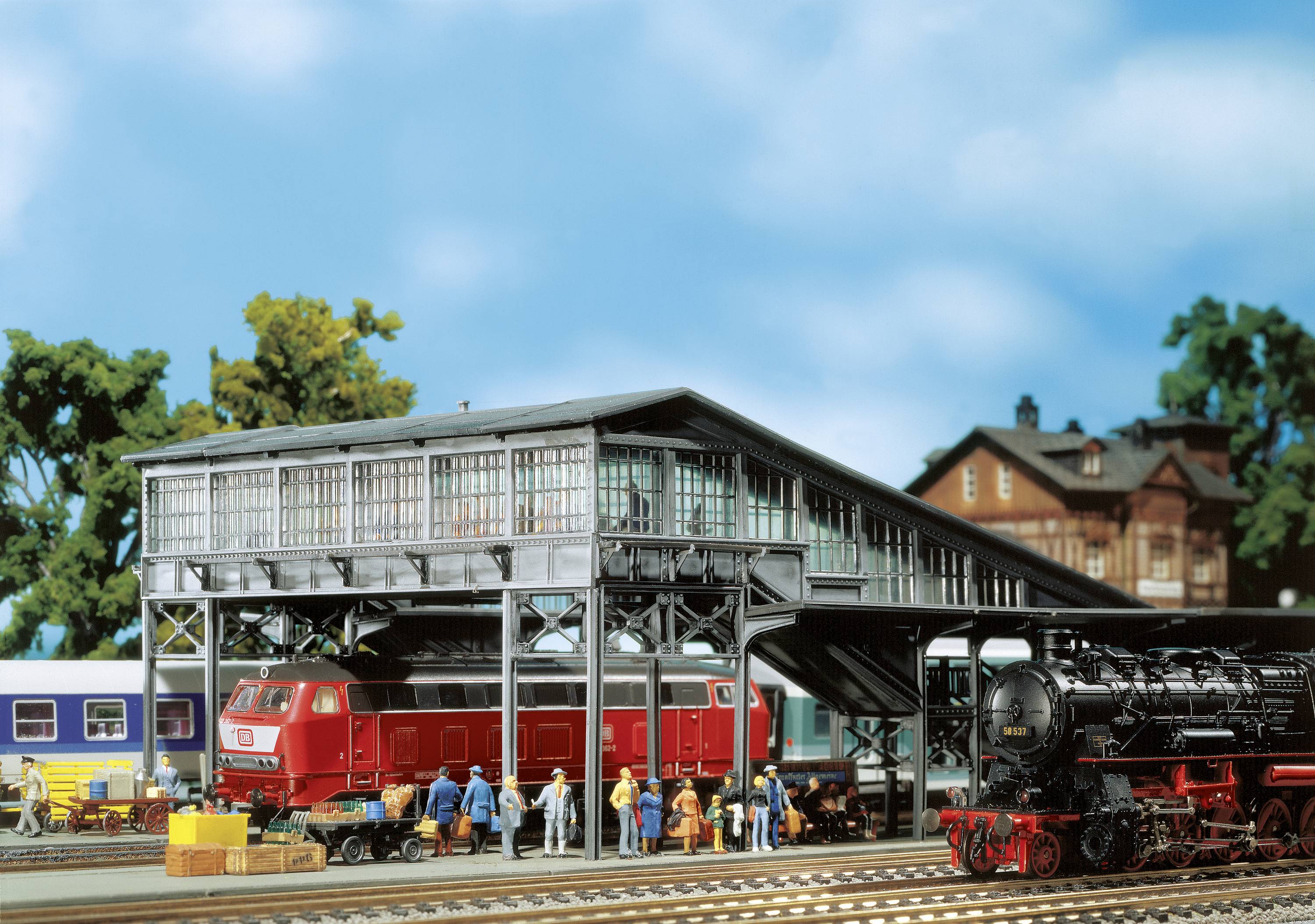 Model železniční scéna s červeným vlakem pod železniční dílnou, obklopená miniaturními figurkami a dalšími vlaky v pozadí.