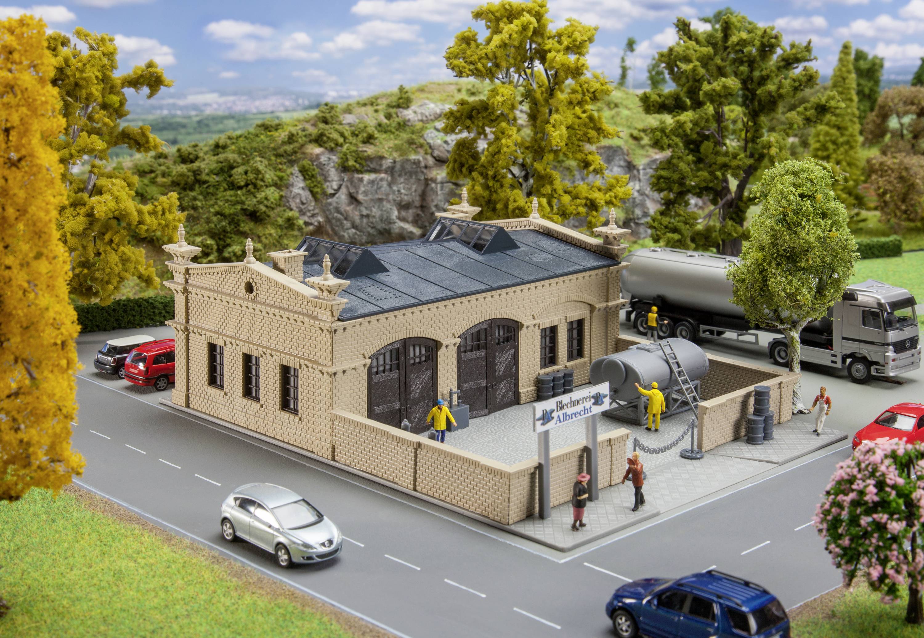 Model historické tovární scény s miniaturními auty a figurkami. Před továrnou je zaparkovaný cisternový vůz.